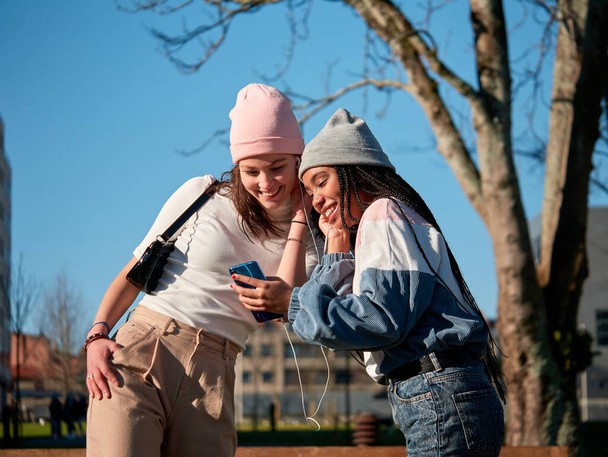 ένα ζευγάρι των όμορφων νεαρών κοριτσιών που χρησιμοποιούν ένα smartphone και ακουστικά, σε εξωτερικούς χώρους, αναζητούν χαρούμενος και φορώντας casual ρούχα - Φωτογραφία, εικόνα