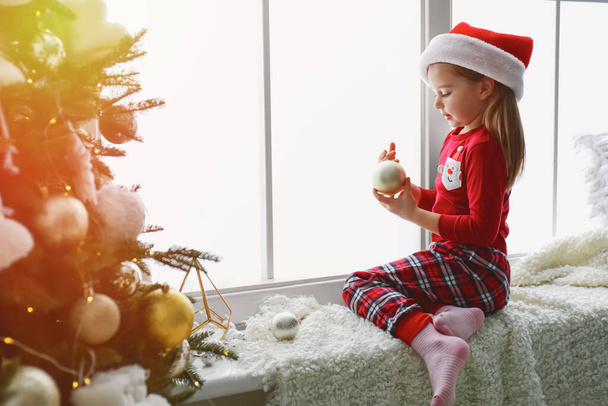 Cute little girl in Santa hat holding Christmas ball on window sill at home - Foto, afbeelding