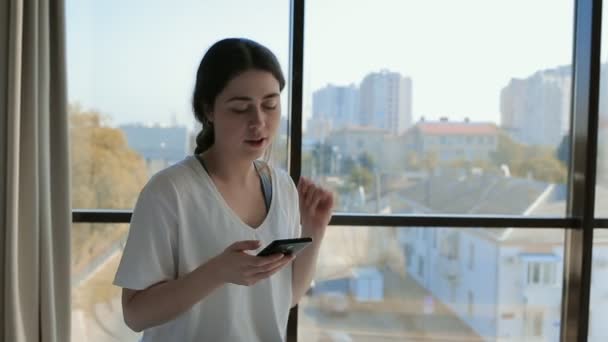 Happiness. Portrait of a young beautiful woman sitting on a bed, listening to music on her smartphone and singing along to songs. Indoors. Windows in the background - Footage, Video