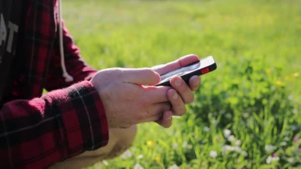 A man uses a smartphone. In the background, bright green grass waving in the wind. Hands close up. Internet and communications - Footage, Video