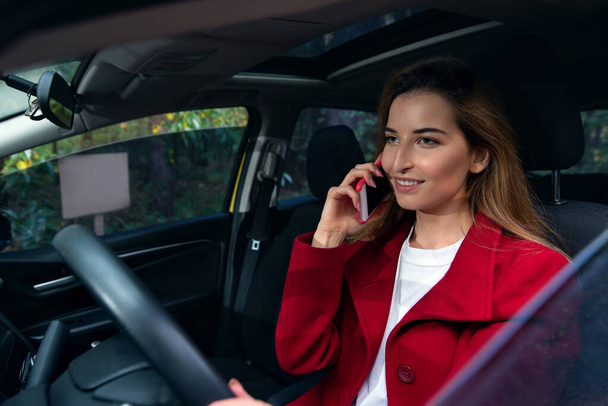 Joven mujer sonriente con un abrigo rojo hablando en el teléfono móvil en el coche en movimiento - Foto, imagen
