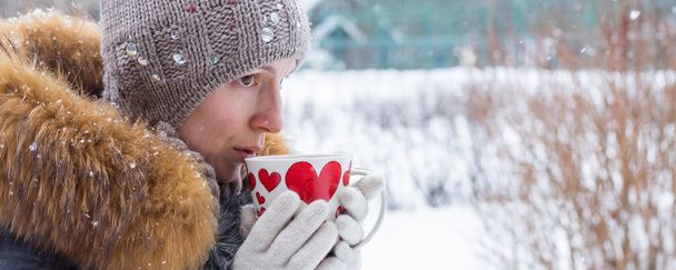 Mujer de mediana edad sosteniendo una taza de bebida caliente en el fondo de una calle de invierno. al aire libre shoot.A chica en chaqueta azul y sombrero de invierno. Ella sostiene una taza de café. invierno, descanso en la ciudad, aperitivos y almuerzo - Foto, Imagen
