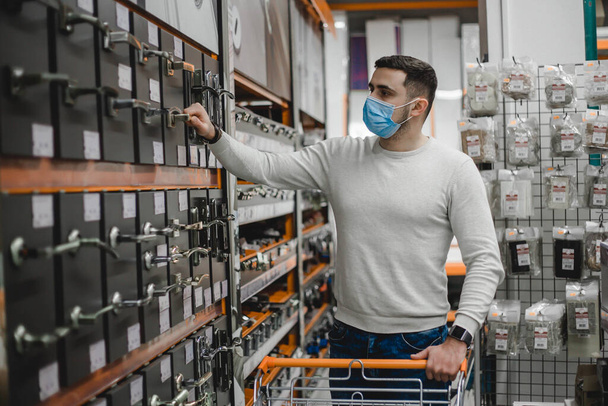 Ritratto di giovane uomo che indossa maschera medica usa e getta scegliendo nuove maniglie delle porte nel negozio di famiglia - Foto, immagini