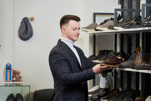 Młody mężczyzna w białym swetrze i garniturze ocenia buty w sklepie odzieżowym. Mężczyzna w butiku z butami. - Zdjęcie, obraz