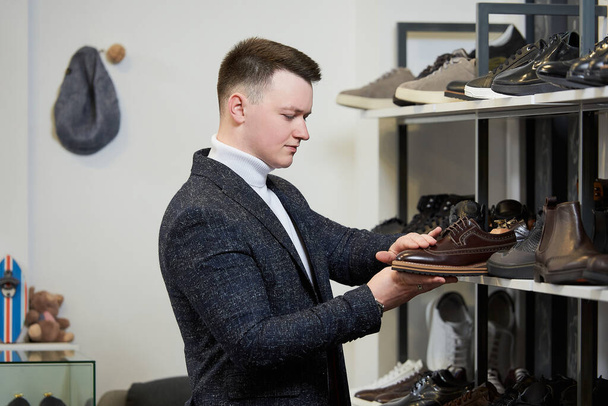 Młody mężczyzna w białym swetrze z golfem i garniturze uważnie ocenia buty w sklepie odzieżowym. Mężczyzna w butiku z butami. - Zdjęcie, obraz