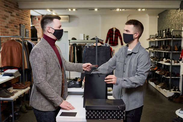顔のマスクの男は、コロナウイルスの拡散を避けるために購入のために支払うために売り手にクレジットカードを渡している。ショップのアシスタントは、ブティックの男性の顧客に服を着た紙袋を与えています。 - 写真・画像