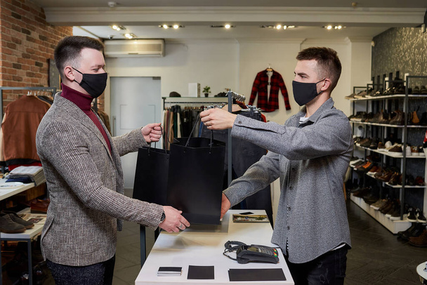Человек в маске для лица, чтобы избежать распространения коронавируса, забирает свою покупку у продавца в магазине одежды. Помощник продавца мужского пола раздает покупателю вторую сумку с одеждой в бутике - Фото, изображение
