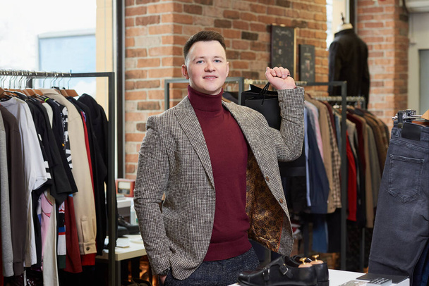 Un joven con una chaqueta deportiva está posando con sus compras en bolsas de papel negro en una tienda de ropa. Un cliente varón feliz en una boutique. - Foto, imagen