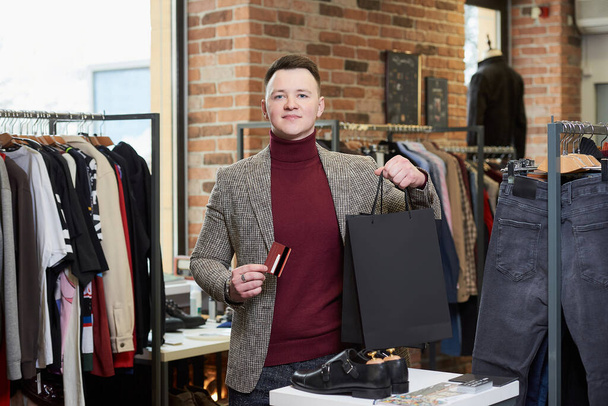 Een jongeman in een sportjasje poseert met zijn aankopen in zwarte papieren tassen en een creditcard in een kledingwinkel. Een gelukkige mannelijke klant in een boetiek. - Foto, afbeelding
