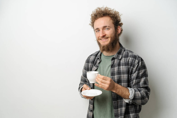 Smiling handsome man with curly hair and beard drinking coffee holding cup, wearing plaid long sleeve shirt isolated on white background - Фото, изображение