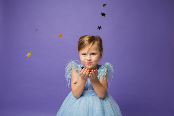 Mavi elbiseli güzel bir kızın portresi mor bir arka planda konfeti yıldızlarını uçuruyor. - Fotoğraf, Görsel