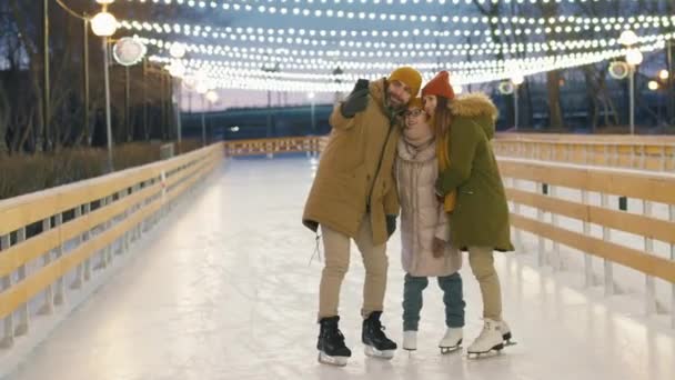 Pieno colpo di gioiosa famiglia caucasica in abbigliamento invernale scattare selfie su smartphone e poi ridere di foto in piedi in mezzo alla pista di pattinaggio vuoto all'aperto - Filmati, video