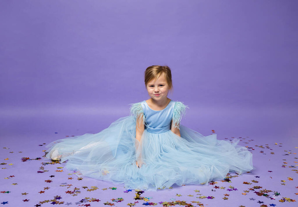 красивая маленькая блондинка в пышном платье принцессы сидит с конфетти на фиолетовом фоне - Фото, изображение