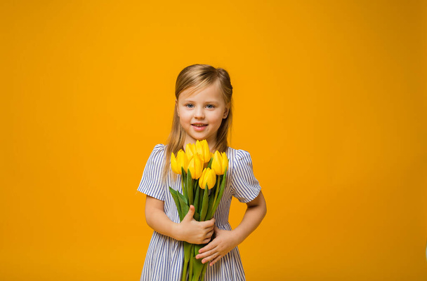Porträt eines schönen kleinen blonden Mädchens in einem gestreiften Kleid mit einem Strauß gelber Tulpen auf gelbem Hintergrund mit Platz für Text - Foto, Bild