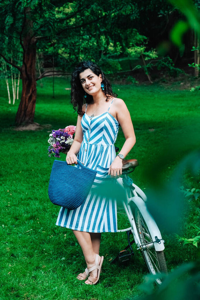 Joven hermosa mujer vestida con un vestido a rayas está de pie junto a una bicicleta con una cesta de flores en el parque. Copiar, espacio vacío para texto. - Foto, imagen