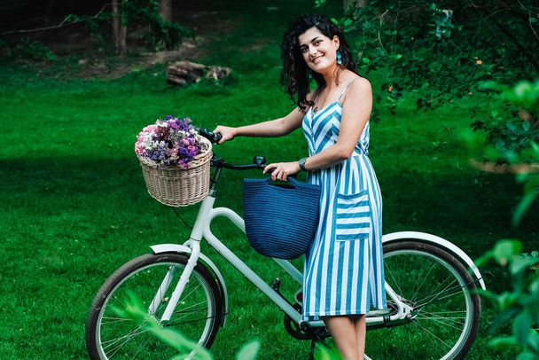 Молода красива жінка, одягнена в смугасту сукню, стоїть поруч з велосипедом з кошиком квітів у парку. Копіювати, порожній простір для тексту
. - Фото, зображення