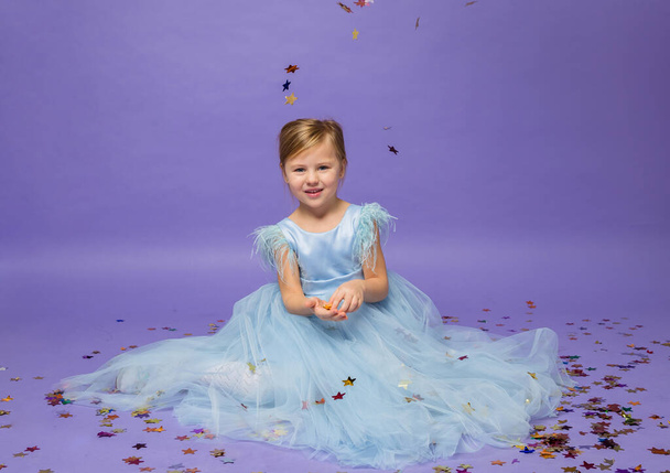 een klein meisje in een full-length blauwe prinses jurk zit op de vloer met confetti op een paarse achtergrond - Foto, afbeelding