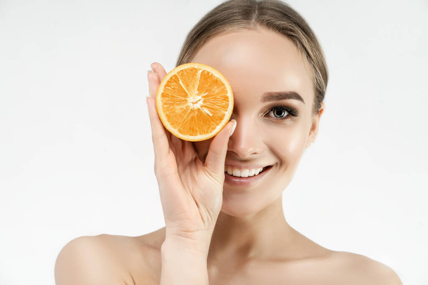 Spa center. Portrait of a beautiful woman covering one eye with an orange. Healthy skin. Blonde woman smiling at the camera. - Foto, afbeelding