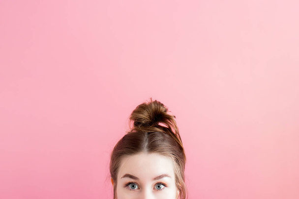 Surprised, amazed young woman with big eyes and hair bun peeking from below, hiding her face and looking at camera over pink background, copy space. Cute millennial girl standing alone. Funny hairdo - Foto, afbeelding