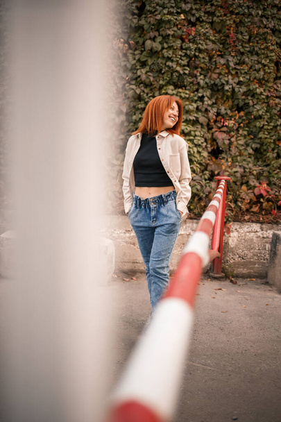 Eine hübsche rothaarige junge Frau läuft die Straße hinunter, sie trägt Jeans und ein beiges Hemd. Schöne Mädchen in lässigem Stil gekleidet mit einem Lächeln im Gesicht - Foto, Bild