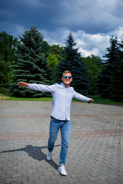 Een knappe jongeman met een Europese uitstraling die een zonnebril draagt, is gekleed in een hemd en jeans. De man loopt de straat op, hij is stijlvol gekleed. - Foto, afbeelding