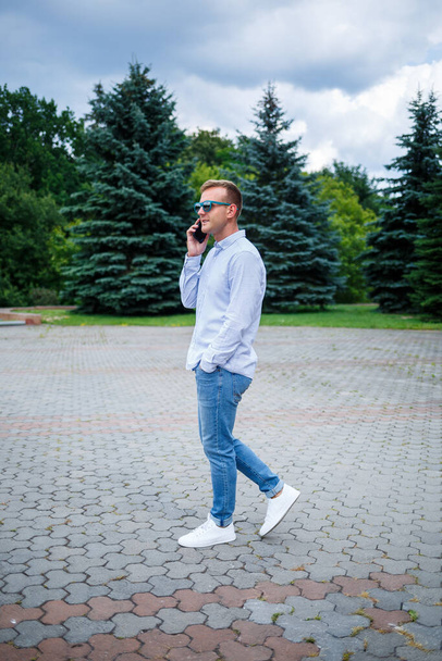A handsome young man of European appearance wearing sunglasses is dressed in a shirt and jeans. The guy walks down the street, he is stylishly dressed - Photo, Image