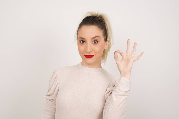 Glad attractive woman shows ok sign with hand as expresses approval, has cheerful expression, being optimistic. Standing against gray wall. - Photo, Image