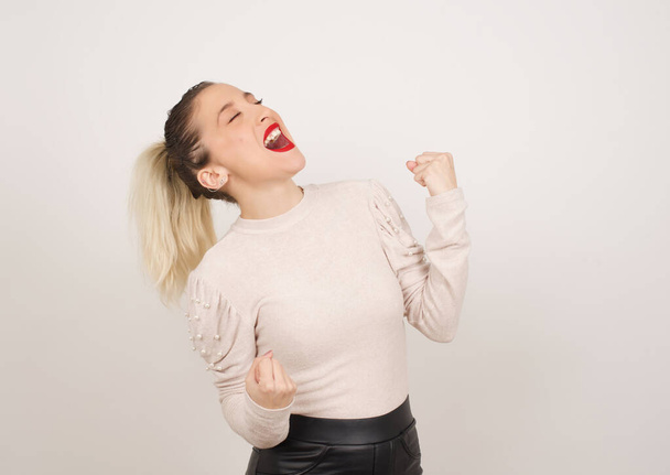 Приваблива молода жінка святкує перемогу, пробиваючи повітря кулаками і блимаючий зубну посмішку над білим студійним фоном з копіювальним простором
. - Фото, зображення