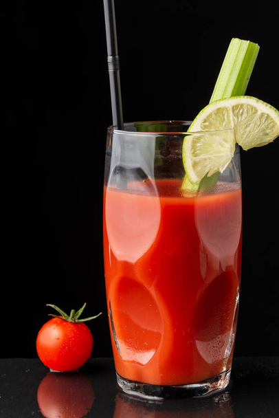 Вид на стакан коктейля Кровавой Мэри с лаймом, сельдереем и соломой, на мокрой доске с помидорами черри, на черном фоне по вертикали, с копировальным пространством - Фото, изображение