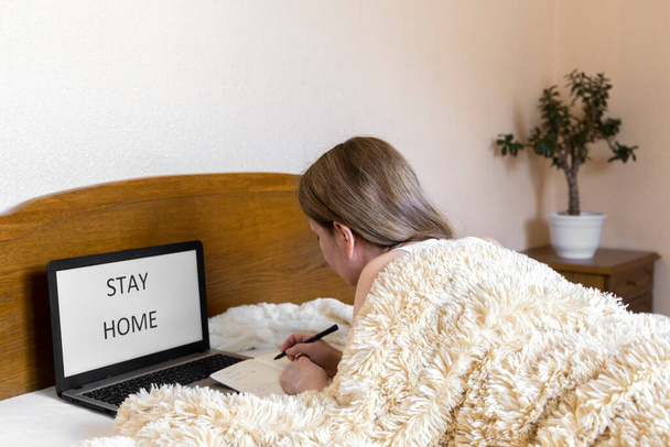 Woman covered with cozy fluffy blanket at home office workplace on the bed with laptop with stay home written on it. Comfortable remote work from home concept - Fotografie, Obrázek