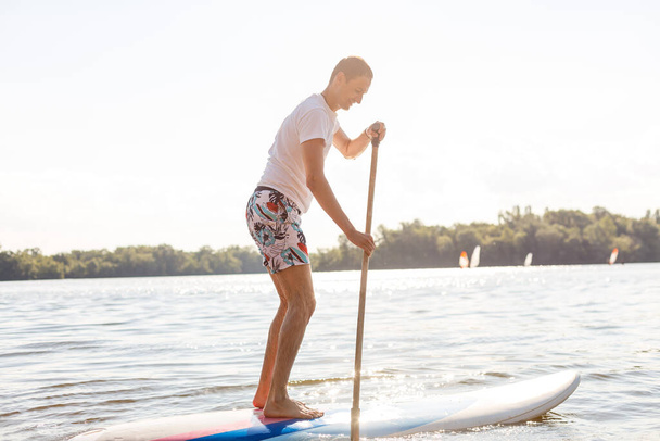 Portrait d'un surfeur avec un SUP Board sur la plage. Jeune homme en planche à pagaie à l'aube. Le concept des sports extrêmes. Style de vie des surfeurs masculins. - Photo, image