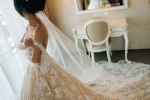 The bride in a beautiful dress with a long train stands near the window in the interior.. - Foto, afbeelding