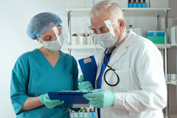 Doctor senior man in witte jas en vrouwelijke paramedicus onderzoeken documenten in blauwe klembord map en bespreken in het ziekenhuis - Foto, afbeelding