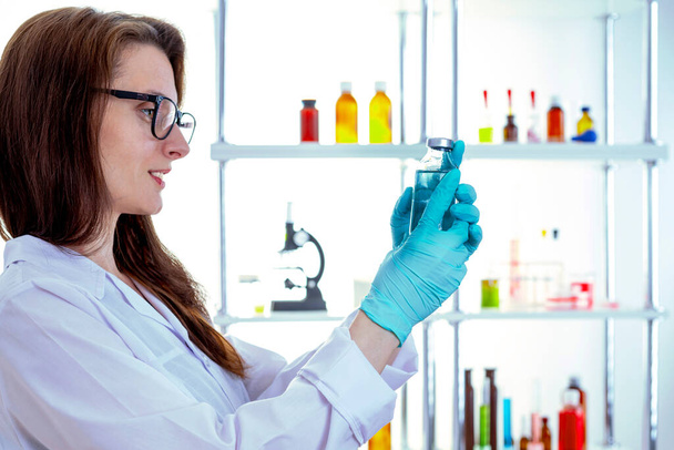 Μια νεαρή χαμογελαστή γυναίκα με ένα λευκό ιατρικό παλτό με γυαλιά εξετάζει ένα μπουκάλι φάρμακα. - Φωτογραφία, εικόνα