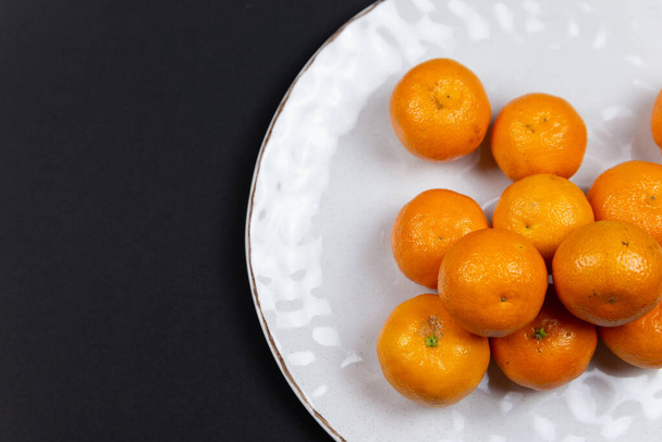 Großer strukturierter Teller mit Mandarinen auf schwarzem Hintergrund. Ansicht der Früchte von oben, mit Platz für Text oder Logo. - Foto, Bild
