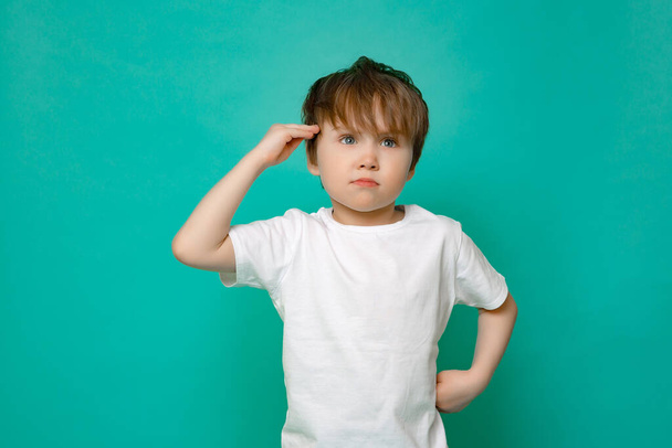 bonito menino de cinco anos de idade em uma t-shirt branca saúda contra um fundo azul isolado. - Foto, Imagem