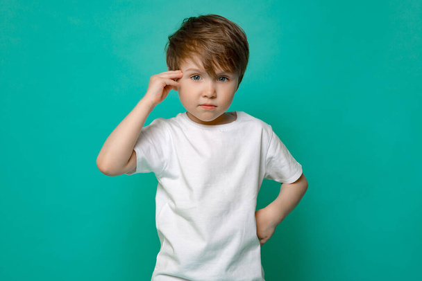 lindo niño de cinco años de edad, en una camiseta blanca saluda contra un fondo azul aislado. - Foto, Imagen