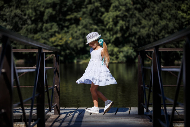 Pieni tyttö, jolla on pitkät hiukset valkoisessa mekossa, seisoo joen rannalla. Kävele järven rannalla aurinkoisena kesäpäivänä. - Valokuva, kuva