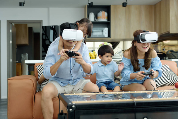 Asyalı bir aile evdeki oturma odasında sanal gerçeklik VR kulaklığı kullanarak oyun oynuyor, neşeli bir aile birlikte eğleniyor. Aile zamanı kavramı - Fotoğraf, Görsel