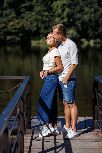Ένα όμορφο ερωτευμένο ζευγάρι στέκεται στην όχθη του ποταμού που αγκαλιάζουν. Ευτυχισμένο παντρεμένο ζευγάρι σε διακοπές δίπλα στη λίμνη - Φωτογραφία, εικόνα