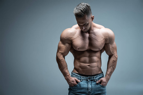 young handsome sportsman bodybuilder posing in jeans on grey background - 写真・画像