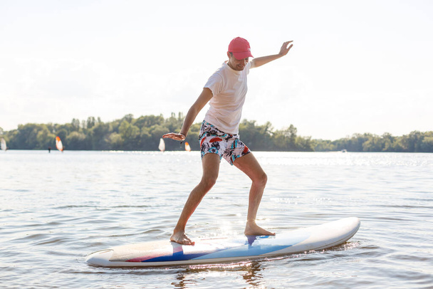Portré egy szörfösről SUP táblával a tengerparton. Fiatalember a táblán hajnalban. Az extrém sport fogalma. Férfi szörfös életmód. - Fotó, kép