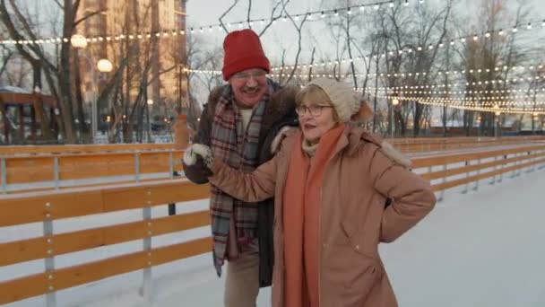 Średnie nagranie szczęśliwej pary starców uśmiechającej się i rozmawiającej podczas jazdy na łyżwach na lodowisku o zachodzie słońca - Materiał filmowy, wideo