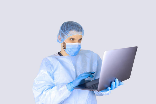 Male doctor in a medical gown, mask and gloves holding a laptop - Fotó, kép