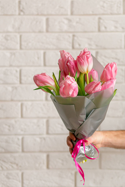Hombre con hermosos tulipanes rosados cerca de la pared de ladrillo blanco. Primer plano con espacio para texto. Tarjeta de felicitación de primavera. Concepto de Día Internacional de la Mujer, Día de la Madre, Pascua. Copiar espacio - Foto, imagen