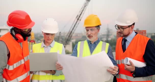 Mixed-races males and female builders with draft plan of building and laptop computer talking on constructing site. Architectors and investor, multi ethnic men and woman constructors working. - Footage, Video