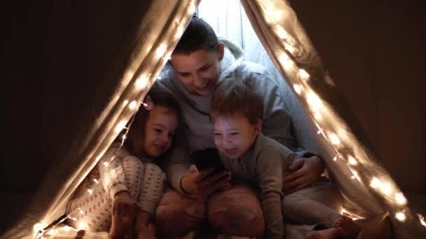 authentic cute mom and preschool kids boy and girl in tent at home. young woman use smartphone with children 2-4 years old in wigwam at night. Family, Childhood, Motherhood, Comfort and Safety concept - Footage, Video