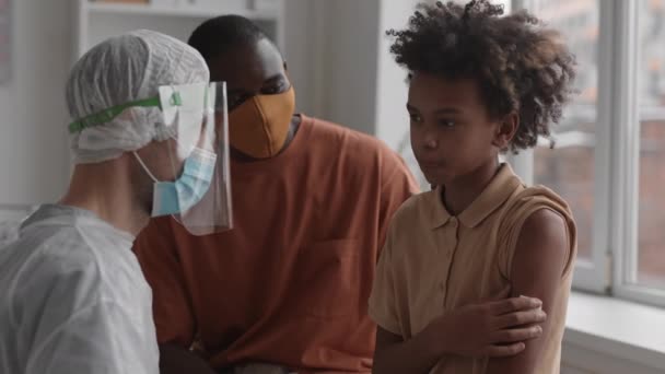 Střední detailní záběr afrického chlapce držícího bavlněnou podložku s rukou na rameni, usmívajícího se, plácajícího nerozpoznatelného lékaře v ochranném obleku poté, co byl očkován, podporujícího bratra sedícího poblíž - Záběry, video