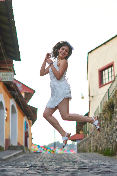 Mujer blanca saltando de felicidad en medio de la calle en sus vacaciones, con vestido blanco y una calle en el fondo - Foto, Imagen