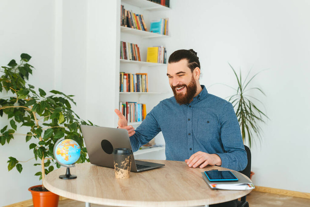 Zdjęcie szczęśliwego przystojnego brodatego mężczyzny czatującego online w biurze - Zdjęcie, obraz